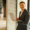 Smiling man in a suit holding a laptop in a modern office hallway.