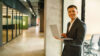 Smiling man in a suit holding a laptop in a modern office hallway.