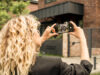 Eine Frau mit blonden Locken fotografiert mit ihrem Smartphone ein modernes Wohnhaus aus Backstein.