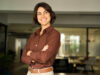 A smiling woman with her arms crossed is standing in a modern office.